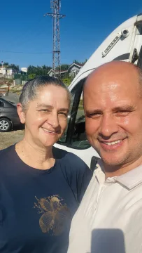 Güneşli bir günde, iki yetişkin açık havada kameraya dönerek yan yana poz veriyor. Arkalarında beyaz bir karavan, kamp aracı veya benzeri büyük bir araç; ayrıca park hâlinde bir otomobil, elektrik direği, kablolar, ağaçlar ve uzakta yapılar görülüyor. Mek