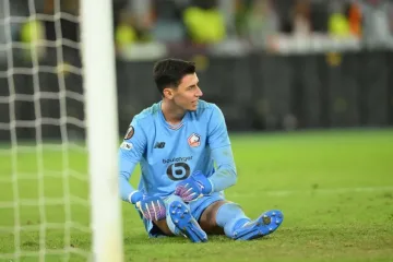 Yeşil çim üzerindeki bir futbol kalesinin önünde oturan genç yetişkin bir kaleci görülüyor. Kişi açık mavi, uzun kollu kaleci forması ve şortu; mavi-beyaz kaleci eldivenleri, açık mavi çoraplar ve mavi kramponlar giyiyor. Kısa, koyu renkli saçları var ve